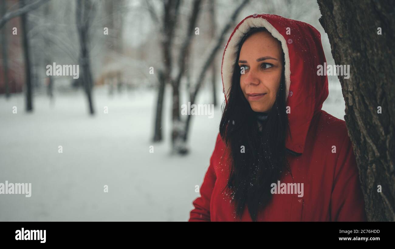 Portrait de la jeune femme en veste rouge et capuche en hiver. Belle dame se tient près de l'arbre et regarde loin. La neige douce enveloppe tout Banque D'Images