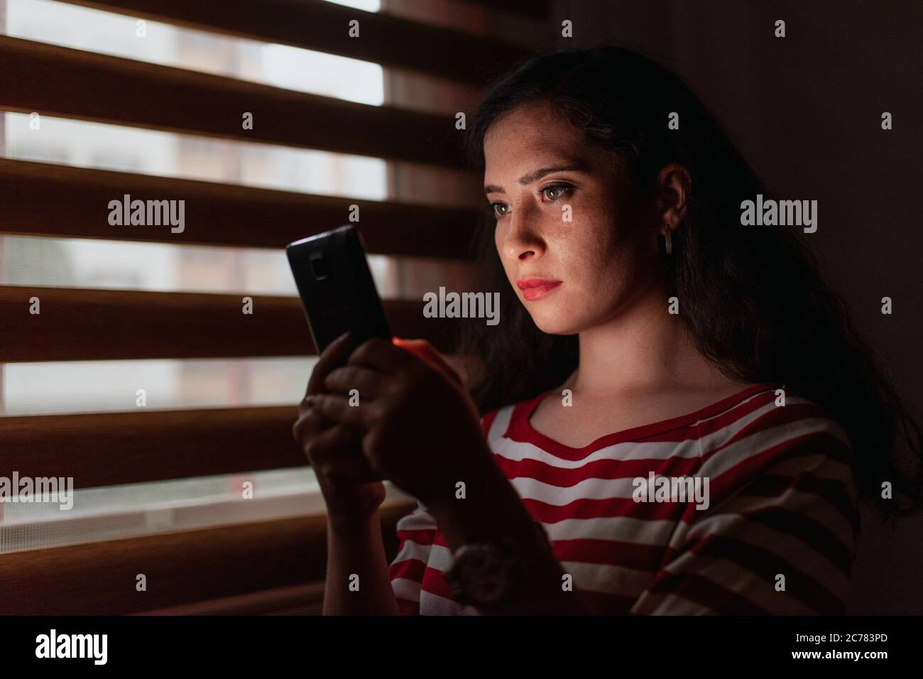 Jeune et sérieuse femme regardant un smartphone dans une pièce sombre Banque D'Images