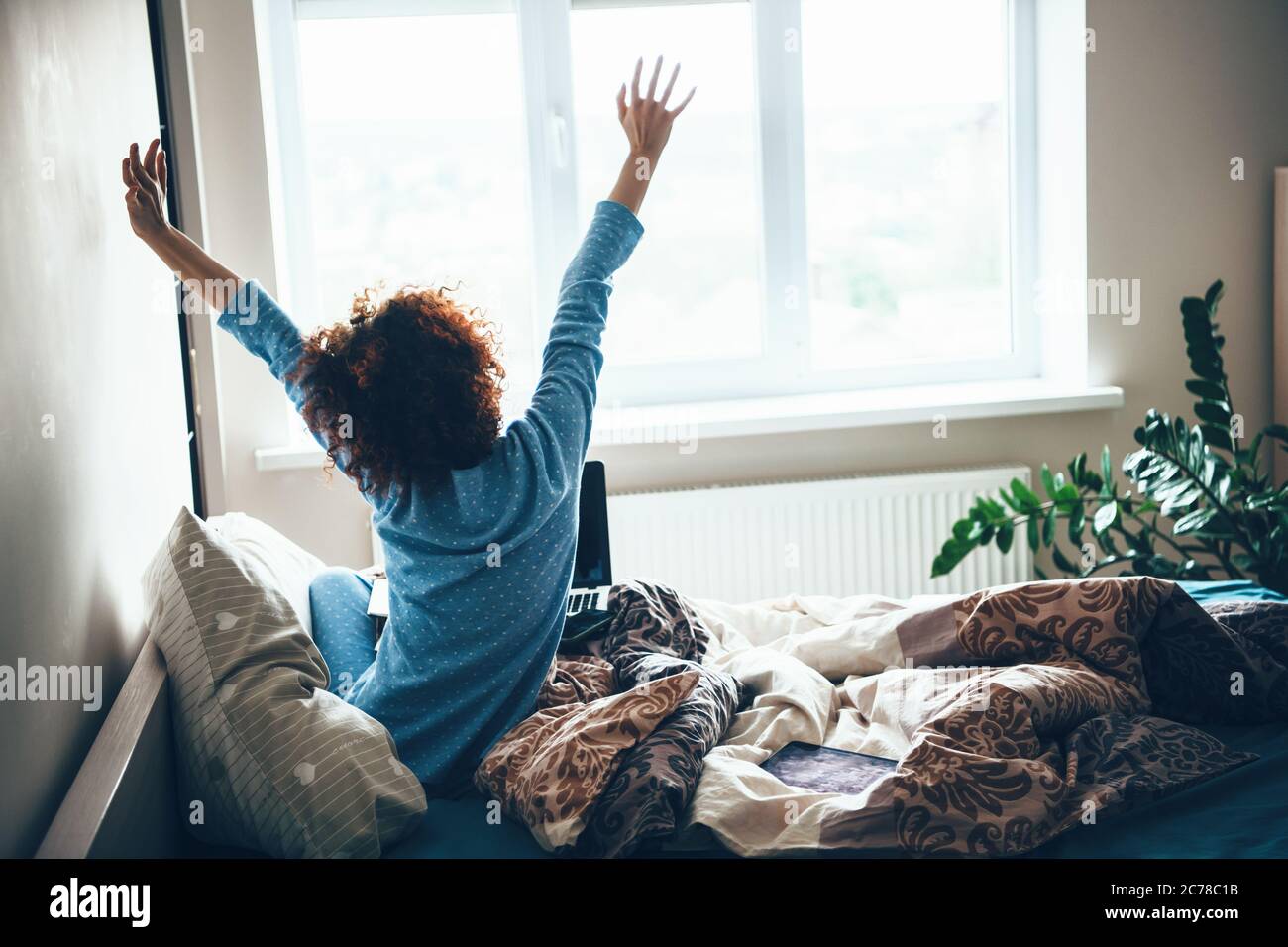 Femme caucasienne aux cheveux bouclés dans un pyjama bleu qui s'étire au lit tôt le matin Banque D'Images