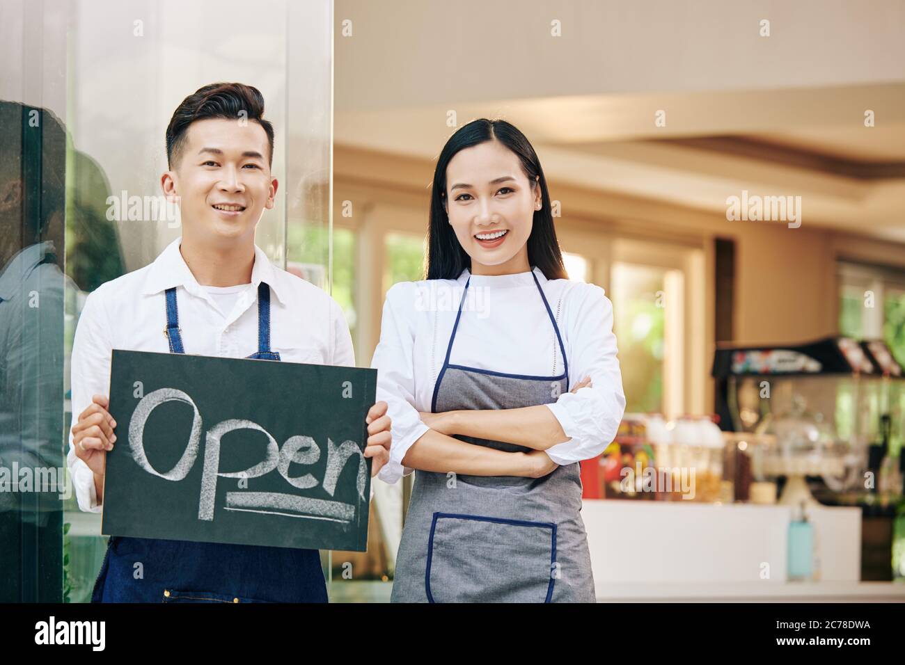 Jeunes propriétaires de cafés vietnamiens souriants avec panneau ouvert à l'entrée du café et accueillant les clients à l'intérieur Banque D'Images