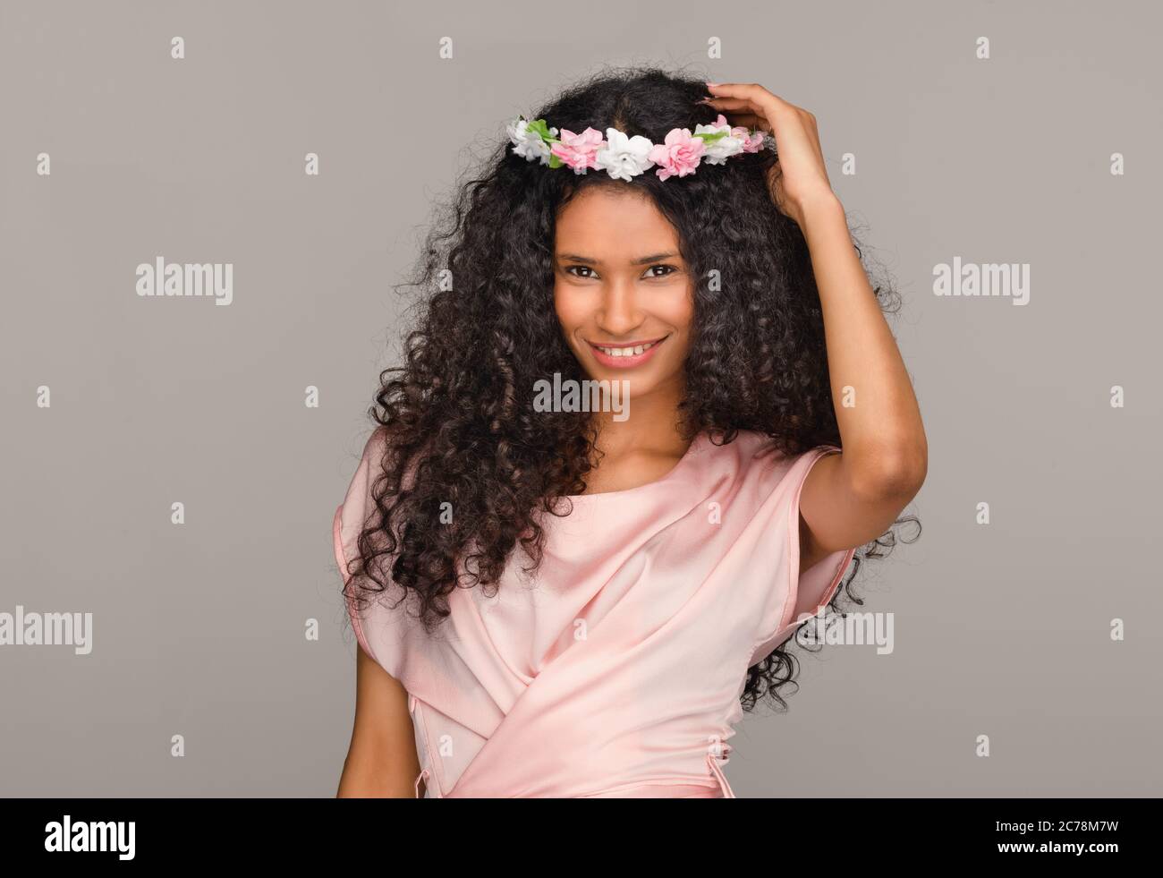 Jolie jeune femme de chambre afro-américaine vêtie d'une robe rose, portant un couronnement de fleurs sur ses cheveux longs et bouclés avec un sourire charmant et joyeux contre un beige Banque D'Images