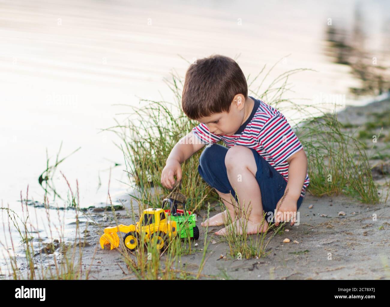 Un petit garçon de 3 ans joue sur la rive de la rivière avec des jouets  lumineux, du sable. Jeux pour enfants près de l'étang en plein air en été  Photo Stock -