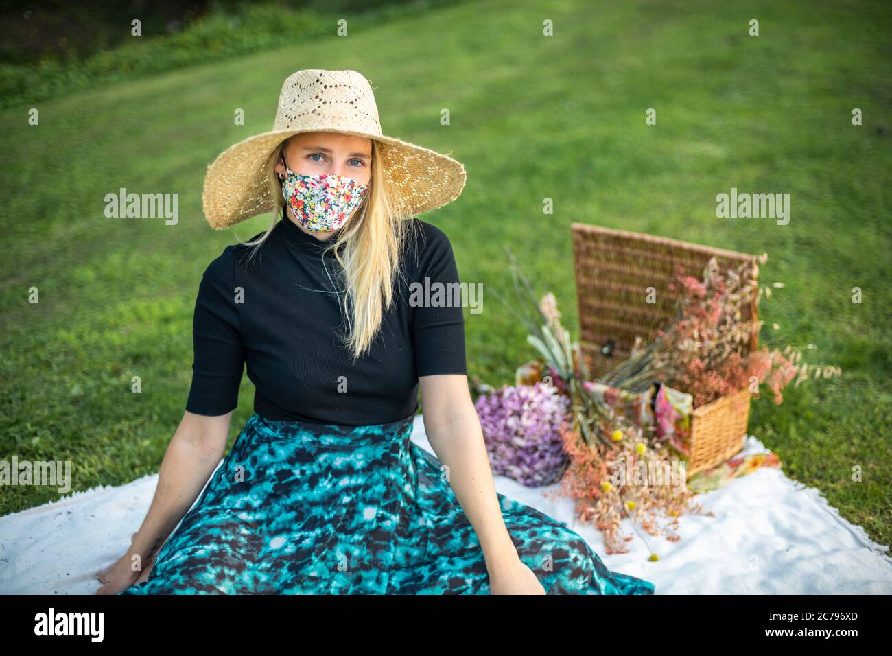 Femme ayant un pique-nique pendant la pandémie de coronavirus Covid-19, portant un masque de visage coloré couvrant dans un champ avec un panier de pique-nique élégant Banque D'Images