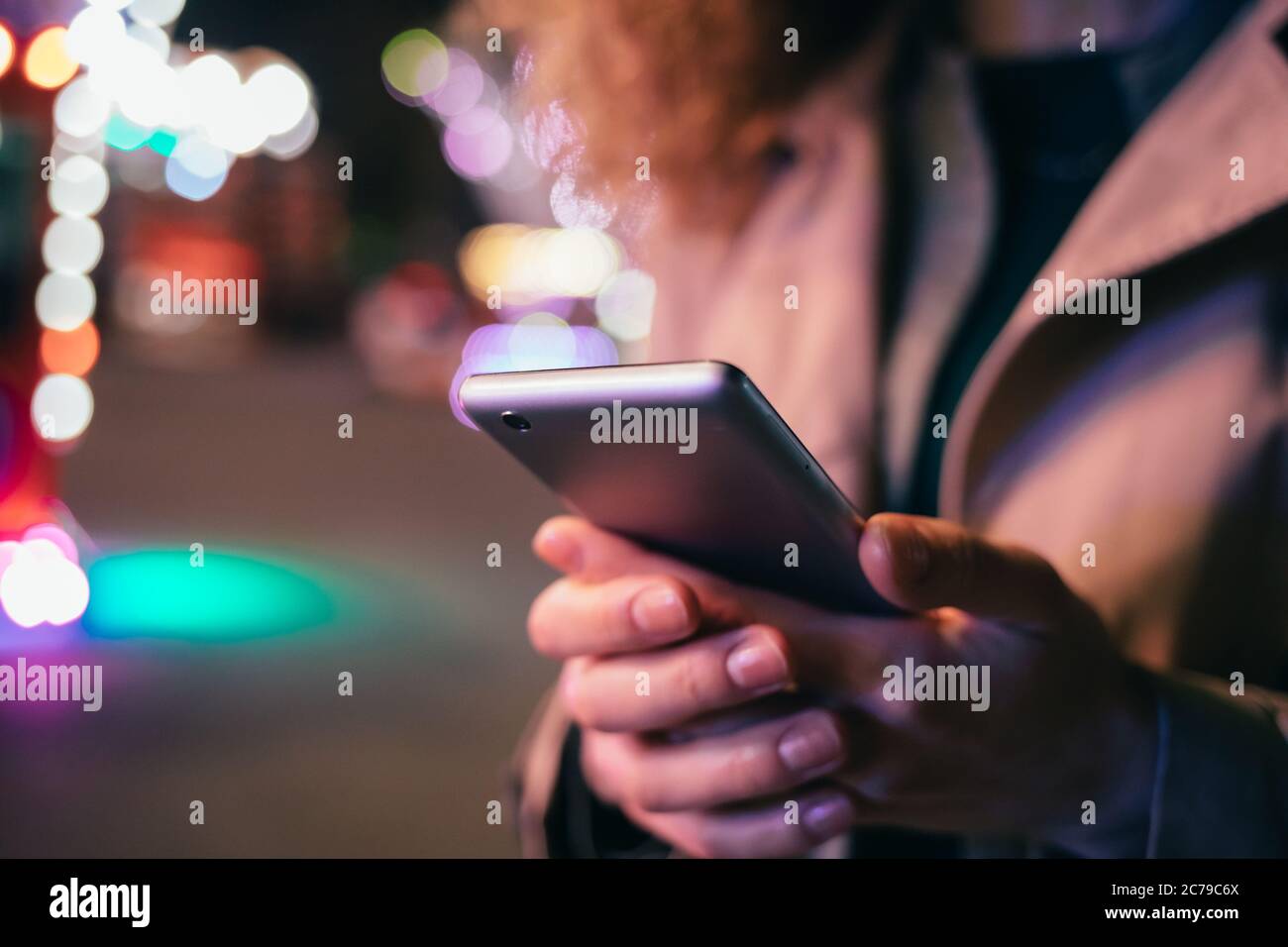 Jeune femme élégante utilisant un téléphone portable debout sur une rue de ville lumineuse de nuit, en gros plan. Banque D'Images