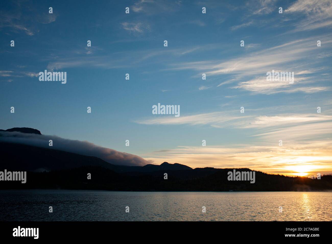 Coucher de soleil doré sur un lac d'Alaska, avec des silhouettes de montagne et des nuages doux reflétant la beauté sereine de la nature sauvage au crépuscule. Banque D'Images