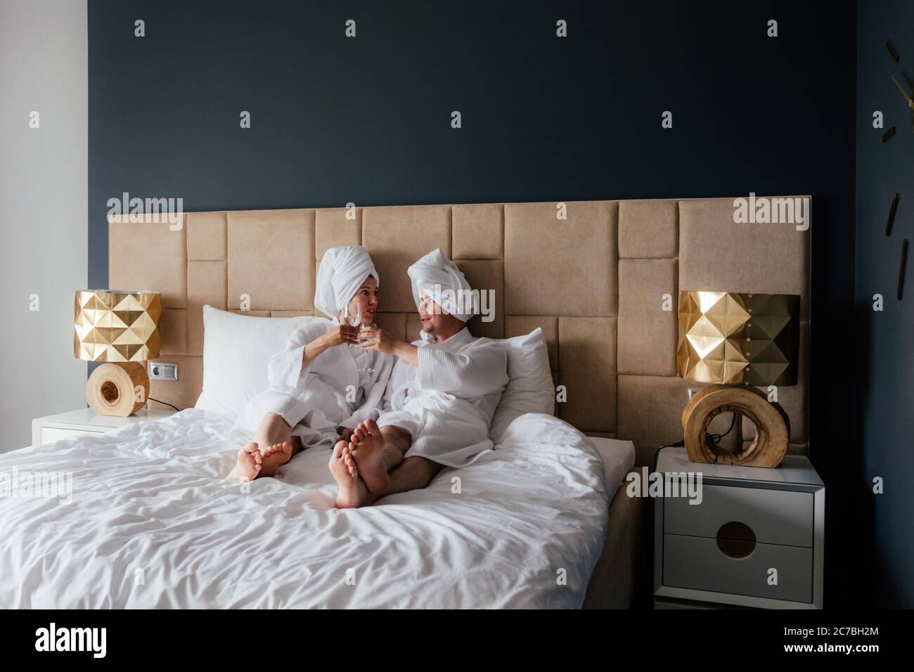 Un jeune couple d'hommes et de femmes en peignoirs blancs et serviettes sur la tête sont couchés dans le lit avec des verres de vin. Banque D'Images