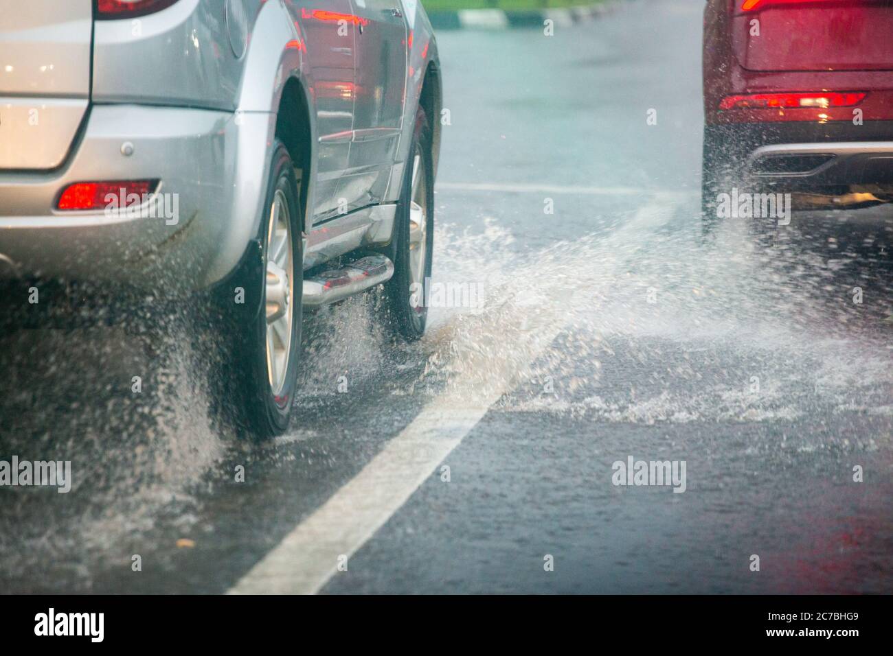 Les éclaboussures d'eau de pluie provenant des roues de voitures argentées et rouges se déplacent rapidement en ville de jour avec une attention sélective. Banque D'Images