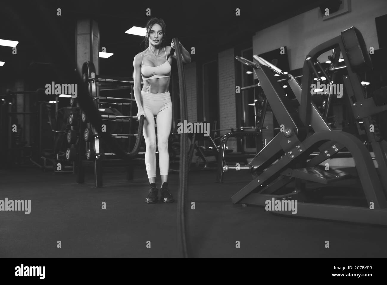 De dessous de vue de la femme brune en sport léger entraînement dans la salle de gym vide avec cordes. Portrait monochrome pleine longueur de la belle fitnesswoman avec le visage fort faisant cardio. Concept de sport. Banque D'Images
