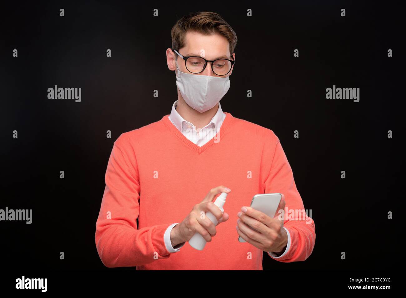 Jeune homme en masque et lunettes désinfectant smartphone avec assainisseur sur fond noir, coronavirus concept Banque D'Images