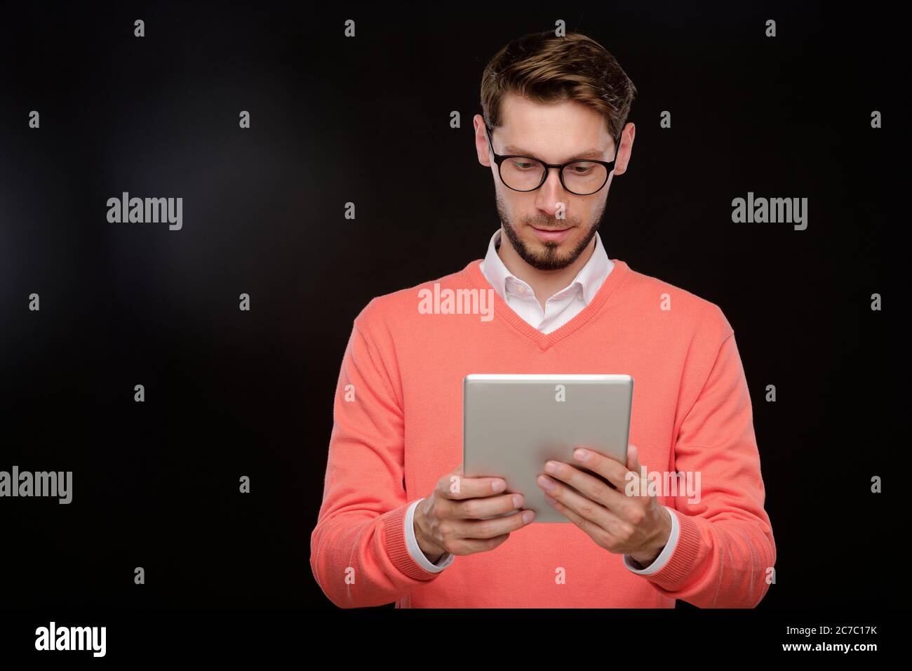 Jeune homme barbu concentré dans un chandail orange utilisant une tablette moderne sur fond noir, concept de médias sociaux Banque D'Images