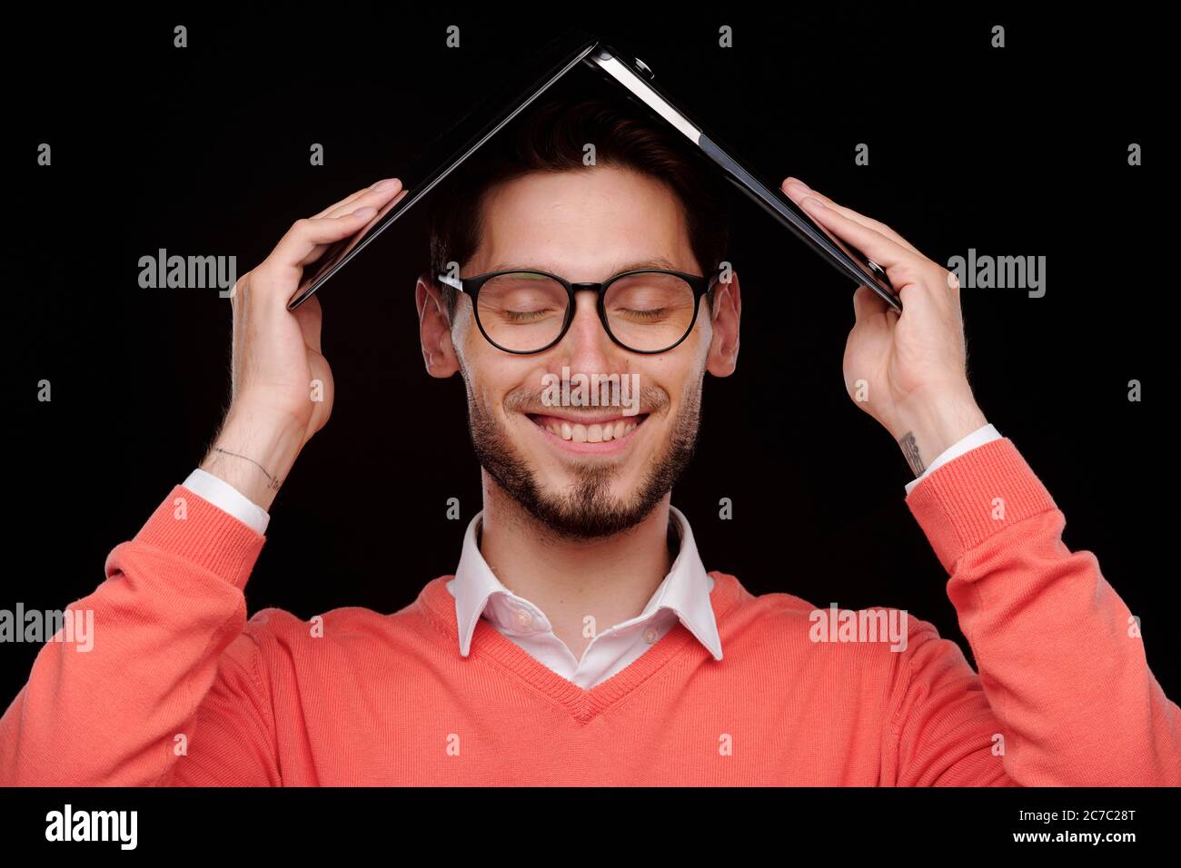 Jeune homme souriant et insouciant, les yeux fermés, gardant l'ordinateur portable au-dessus de la tête tout en se cachant du travail Banque D'Images