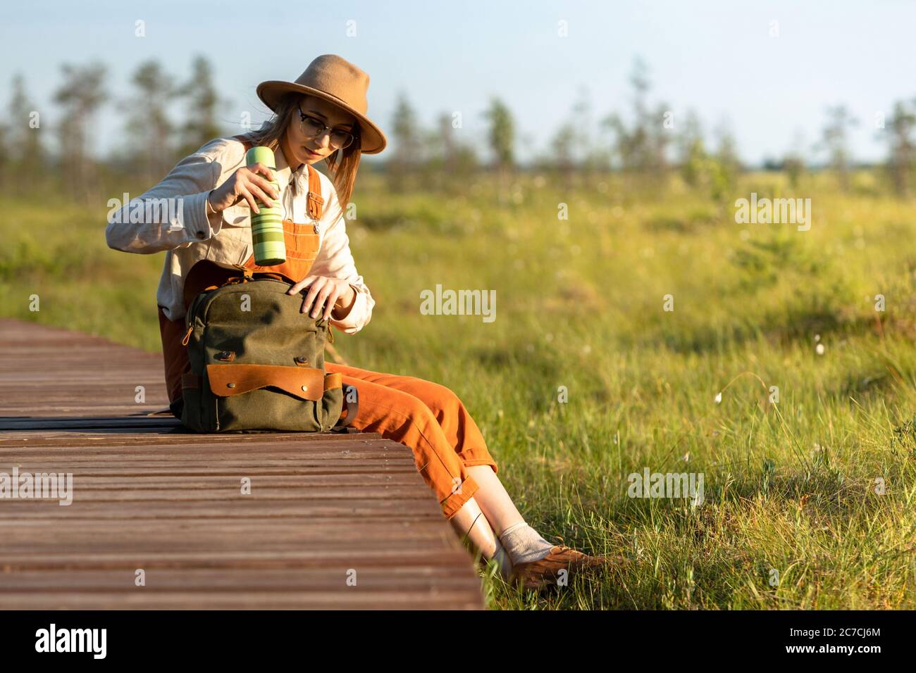 Jeune femme botaniste dans un chapeau assis sur un chemin en bois à travers  le marais de tourbière dans le parc national de la faune.naturaliste  reposant sur la promenade, prend un thermos