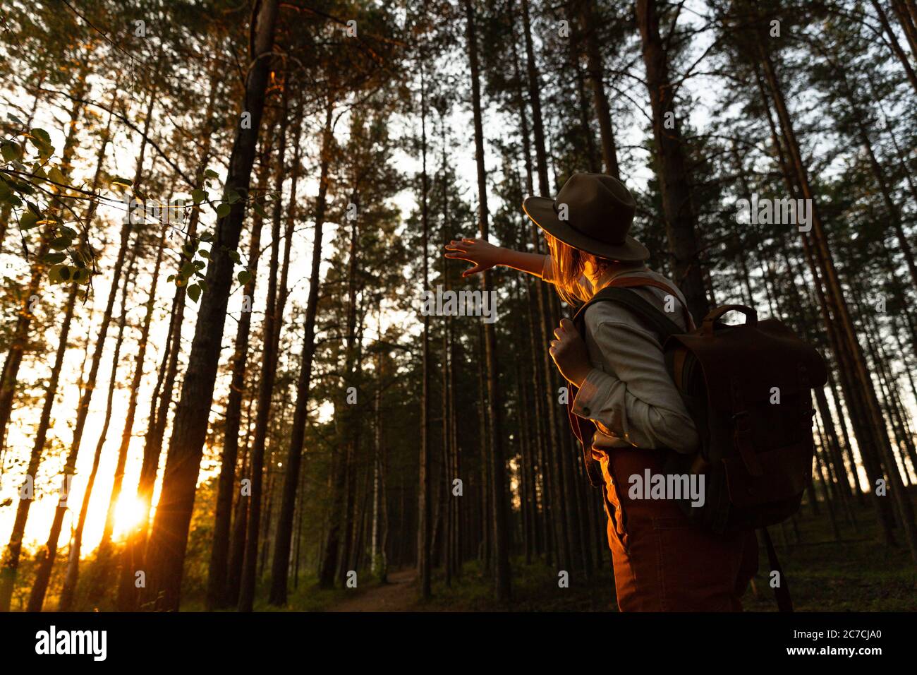 Femme naturaliste en chapeau et combinaison d'orange avec sac à dos regardant le soleil à travers ses doigts au coucher du soleil, explorant la faune et l'écotourisme aventur Banque D'Images