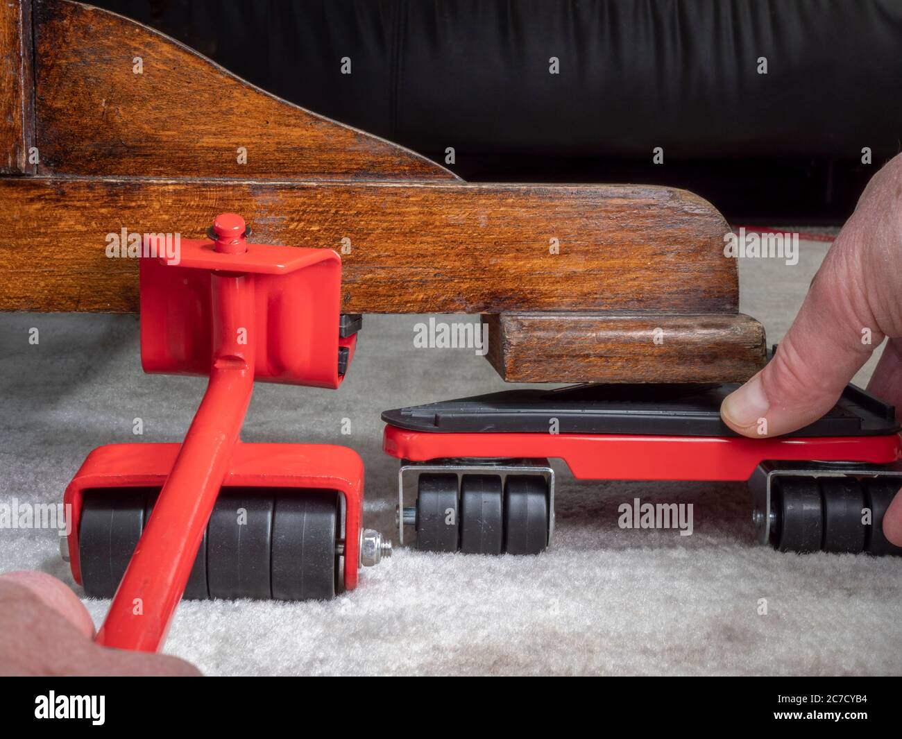 Gros plan de la vue subjective des mains d'un homme à l'aide d'un outil de levage / déplacement de meubles sur un coin de table, avec une plaque de roulettes pour prendre le poids avant de l'abaisser. Banque D'Images