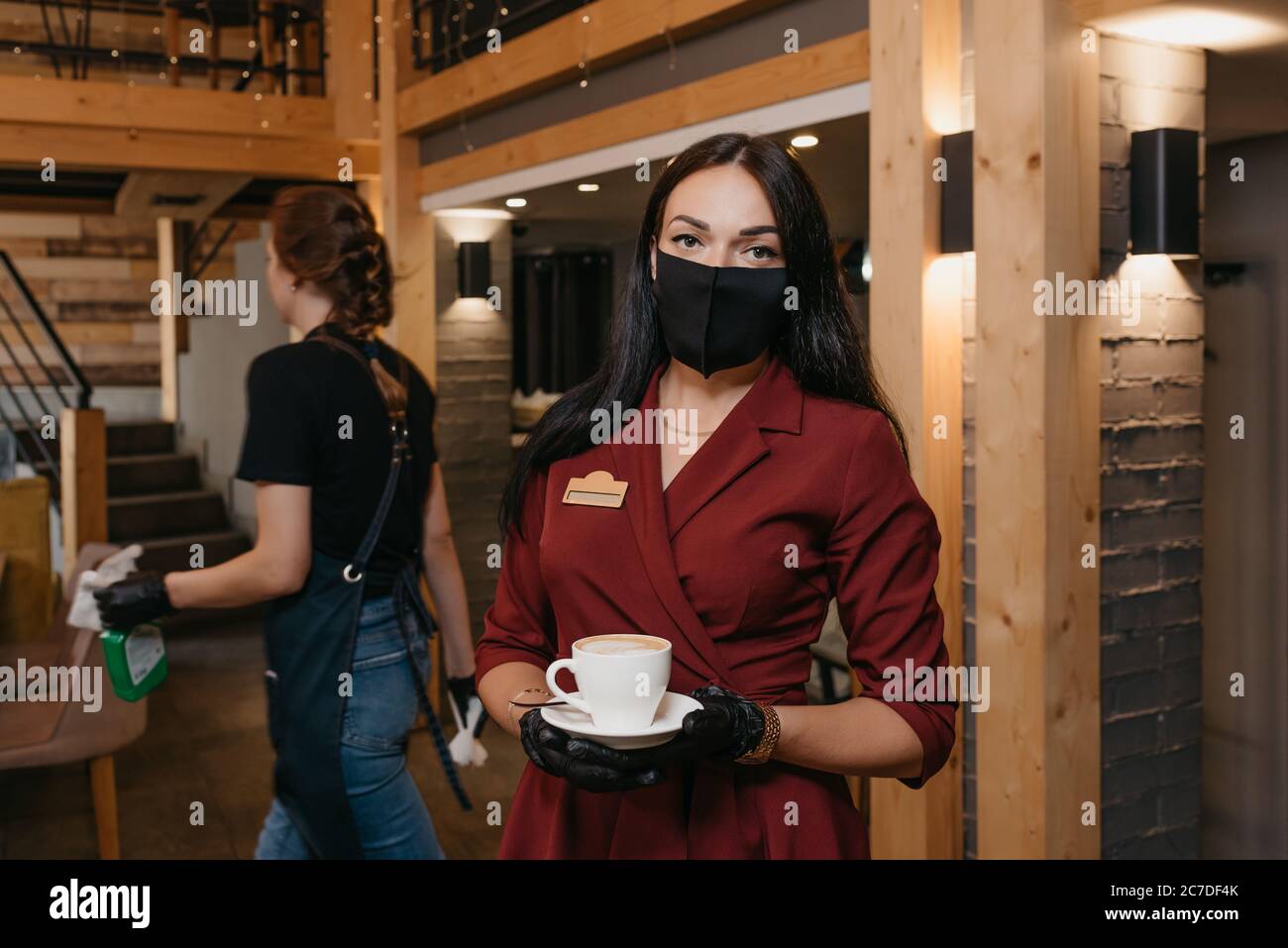 Une magnifique directrice de restaurant qui porte un masque noir et des gants jetables tient une tasse de café dans un restaurant. Une serveuse est propre Banque D'Images