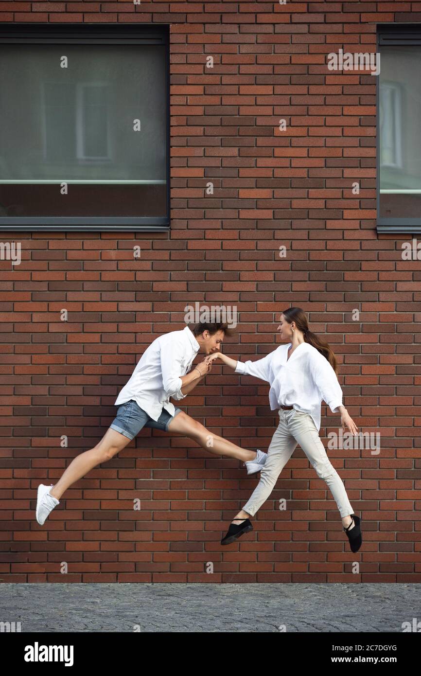 Embrasser, saluer. Saut jeune couple devant le mur de la construction de la ville, sur la course en saut haut. Se hante, se déplace vers la routine quotidienne inspiré et sportif. Jeunes danseurs de ballet vêtus de vêtements décontractés. Banque D'Images