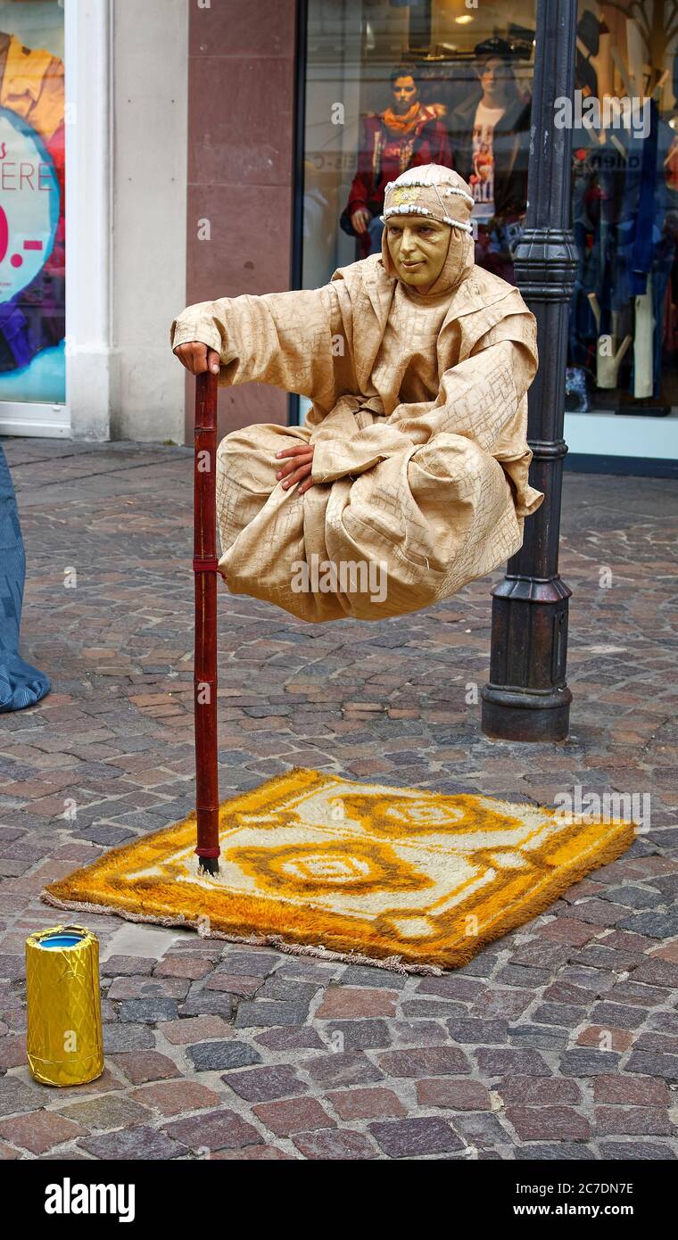 mime mâle, suspendue au-dessus du trottoir; tapis sous, visage peint en or, robe bronzante, intelligent; gagner de l'argent; travail, imaginatif, rue commerçante, pavés, Banque D'Images