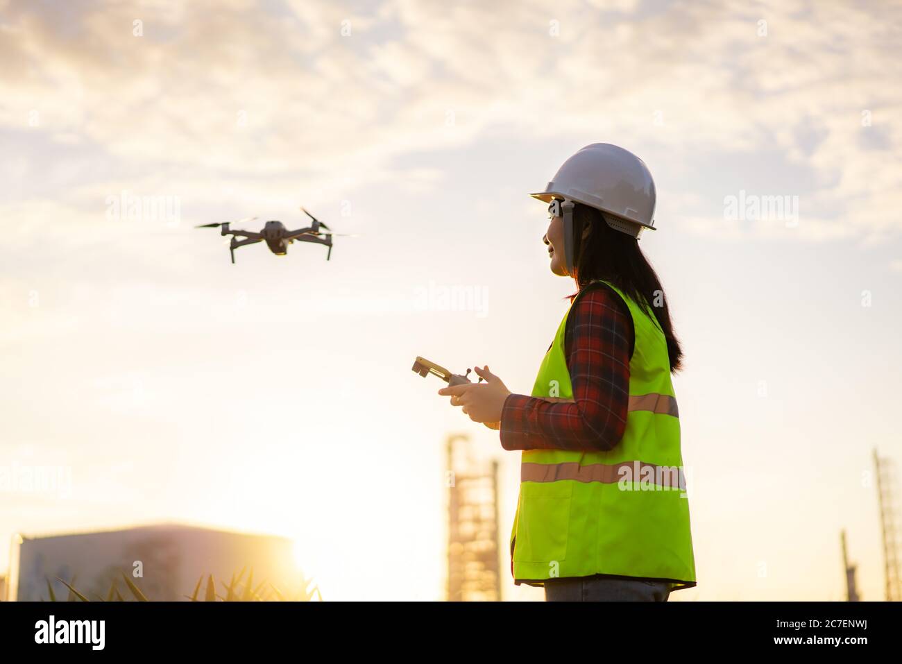 Une femme ingénieur asiatique exploite un drone volant au-dessus de l'usine de raffinerie de pétrole lors d'une étude de chantier de construction au lever du soleil dans le cadre d'un projet de génie civil. Banque D'Images