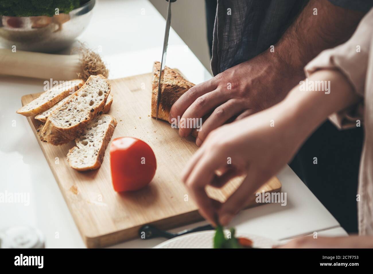 Homme caucasien tranchant du pain pendant que sa femme prépare une salade de légumes dans la cuisine Banque D'Images