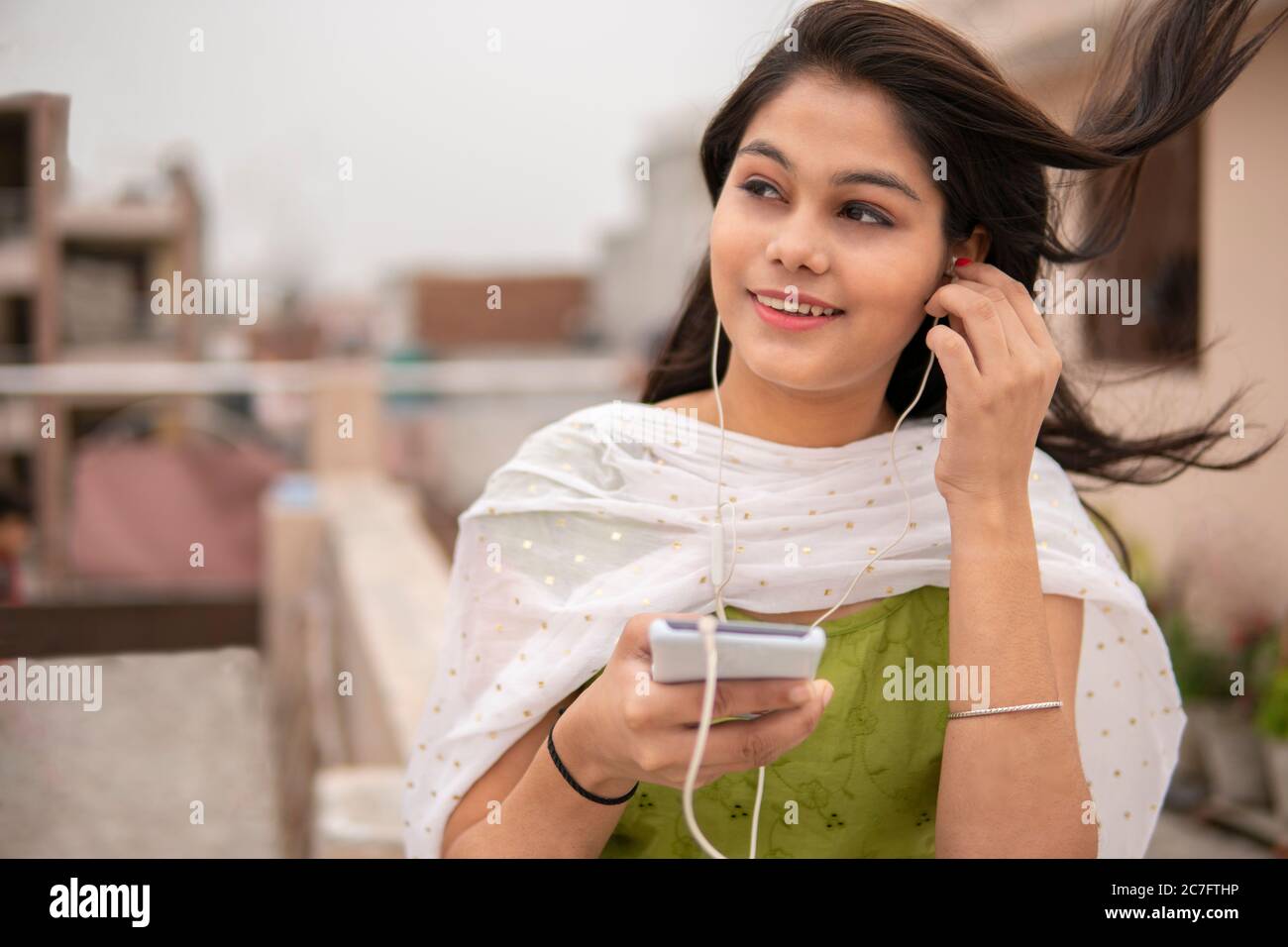 Belle, heureuse jeune fille indienne tard l'écoute de la musique sur smartphone à travers des écouteurs et profiter de l'air frais à l'extérieur sur le toit à l'heure de la journée. Banque D'Images