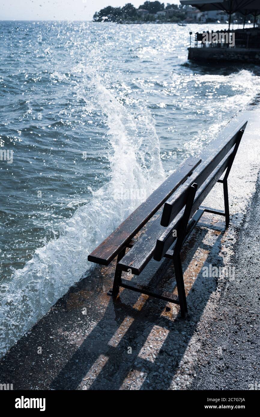 Un banc de parc sombre se dresse sur un quai où les vagues se brisent et les jets d'eau sautent au soleil avec le ciel bleu et la mer. Banque D'Images