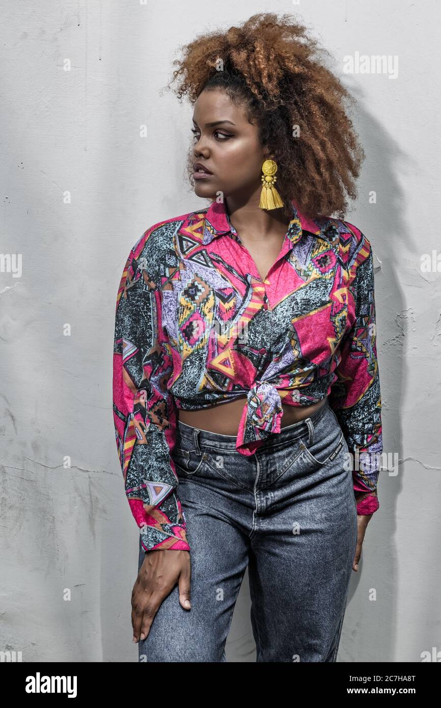 Jeune femme afro-américaine tendance avec coiffure afro et boucles  d'oreilles à pampilles jaunes portant une chemise tendance colorée avec  noeud et un Jean élégant sta Photo Stock - Alamy