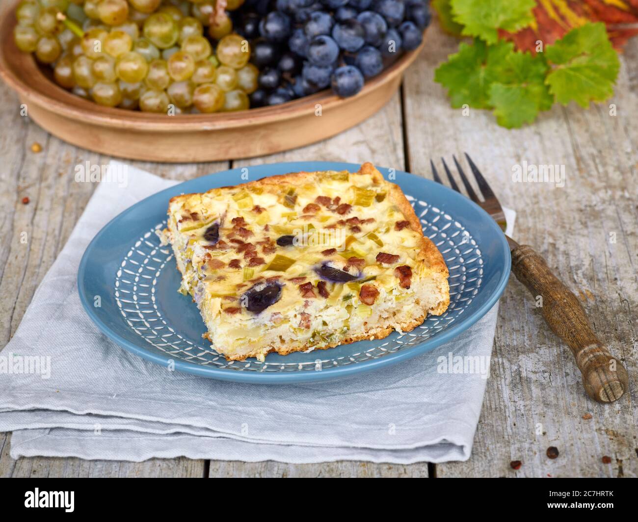 Quiche aux raisins et poireaux, assiette bleue sur toile de lin, raisins dans un bol, fourchette avec manche en bois, table avec plan de table non traité Banque D'Images