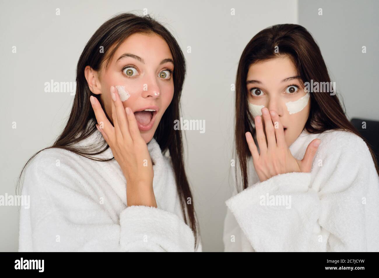 Deux jeunes femmes attrayantes avec des cheveux foncés dans des peignoirs blancs, assis sur le lit avec un masque cosmétique sur les visages, regardant avec étonnement dans l'appareil photo de l'hôtel Banque D'Images