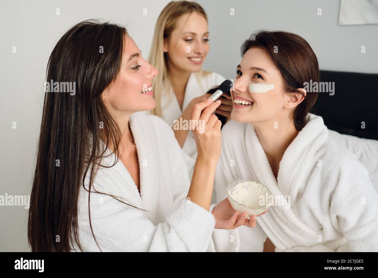 Trois jeunes femmes magnifiques dans des peignoirs blancs assis sur le lit font heureusement un masque cosmétique ensemble dans un hôtel moderne Banque D'Images