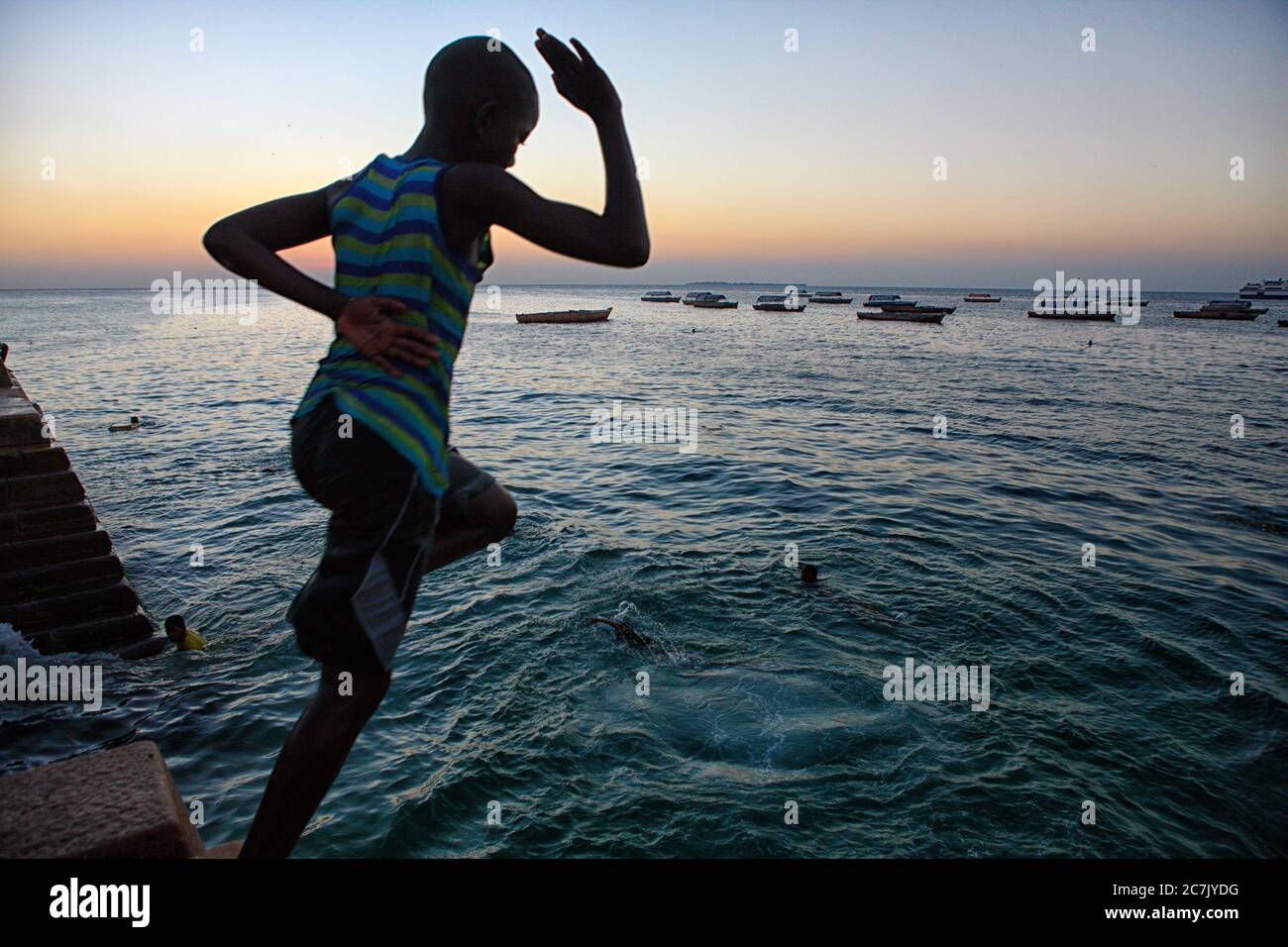 Un garçon non identifié saute dans la mer comme le soleil descend dans la vieille ville en pierre de Zanzibar, Zanzibar, Tanzanie, l'île a une longue histoire avec le commerce des esclaves et a été dirigé par les Arabes omanais au début du XIXe siècle, L'île est majoritairement musulmane et est devenue une destination touristique populaire ces dernières années, l'île est une promenade en bateau de deux heures de Dar es Salaam, Tanzanie sur la côte est africaine, l'île fait partie de la Tanzanie, Banque D'Images