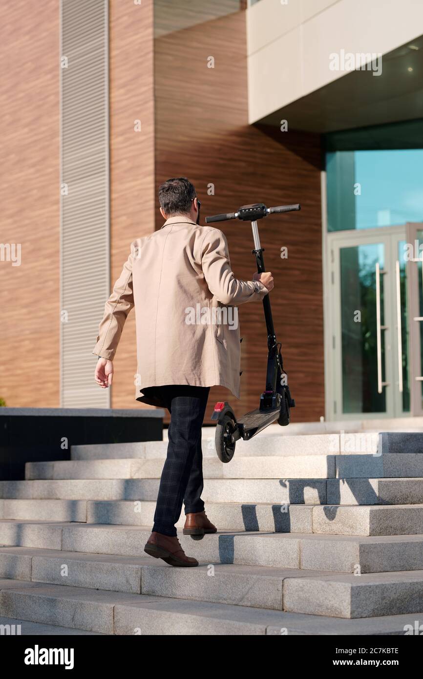 Vue arrière de jeune homme d'affaires élégant en vêtements décontractés élégants portant un scooter électrique en allant à l'étage pour entrer dans le centre de bureau Banque D'Images