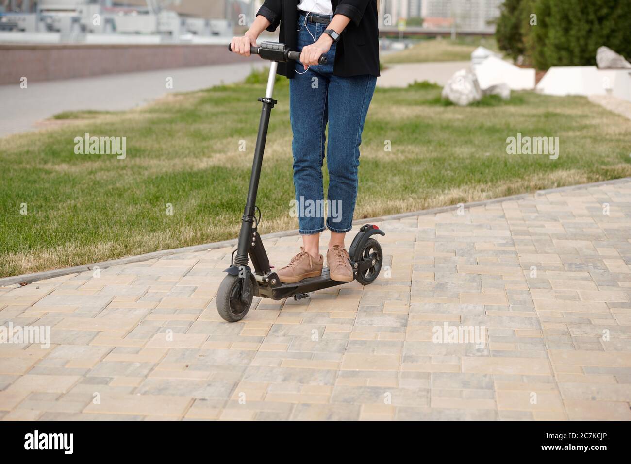 Les jambes et les mains de jeunes femmes contemporaines en vêtements décontractés debout sur un scooter électrique tout en descendant la route urbaine pour travailler Banque D'Images