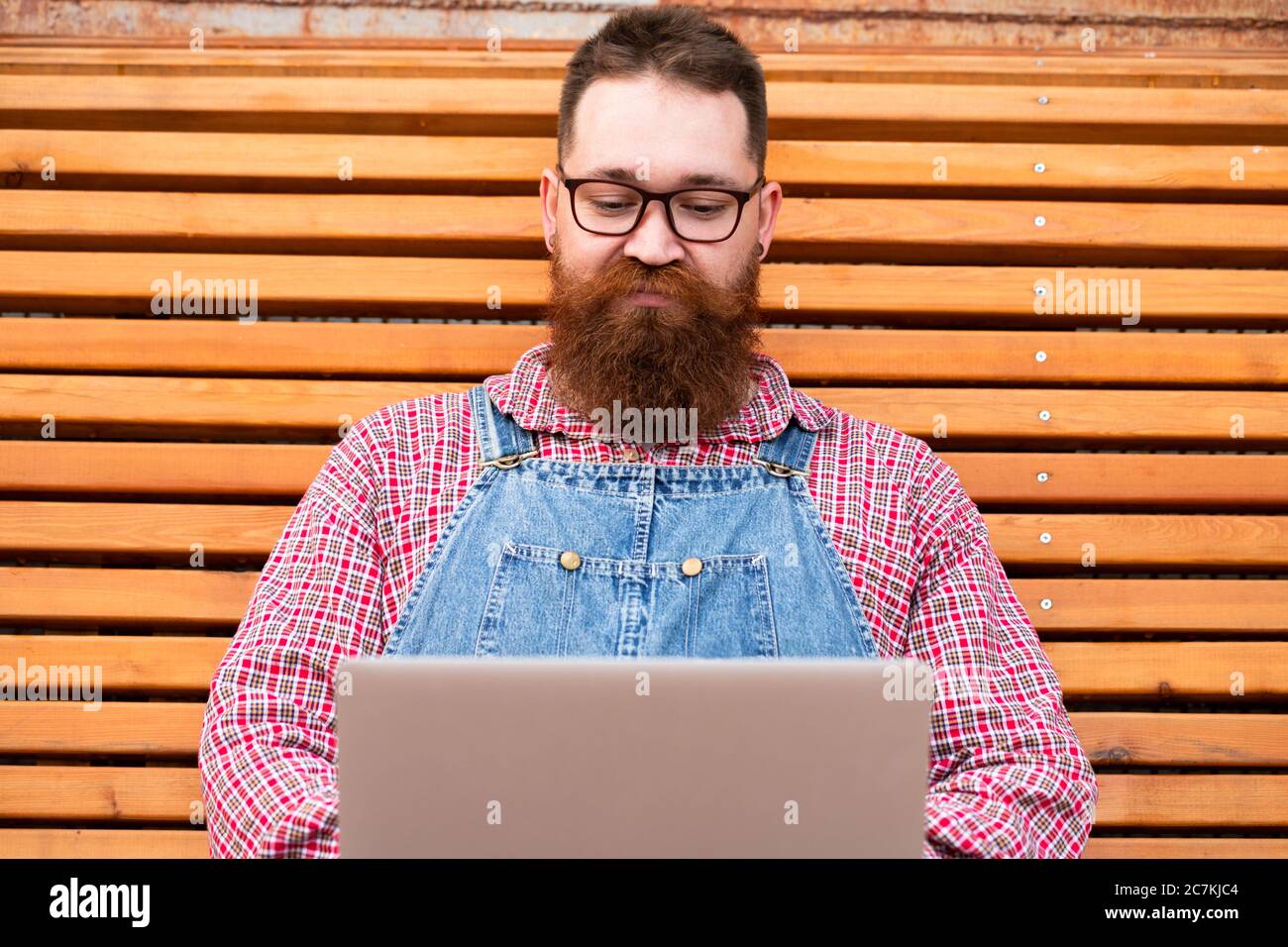 Gros plan portrait d'un boxeur taille basse brutal en combinaison bleue, chemise à carreaux travaillant sur un ordinateur portable assis sur un banc à l'extérieur. Travail à distance Banque D'Images