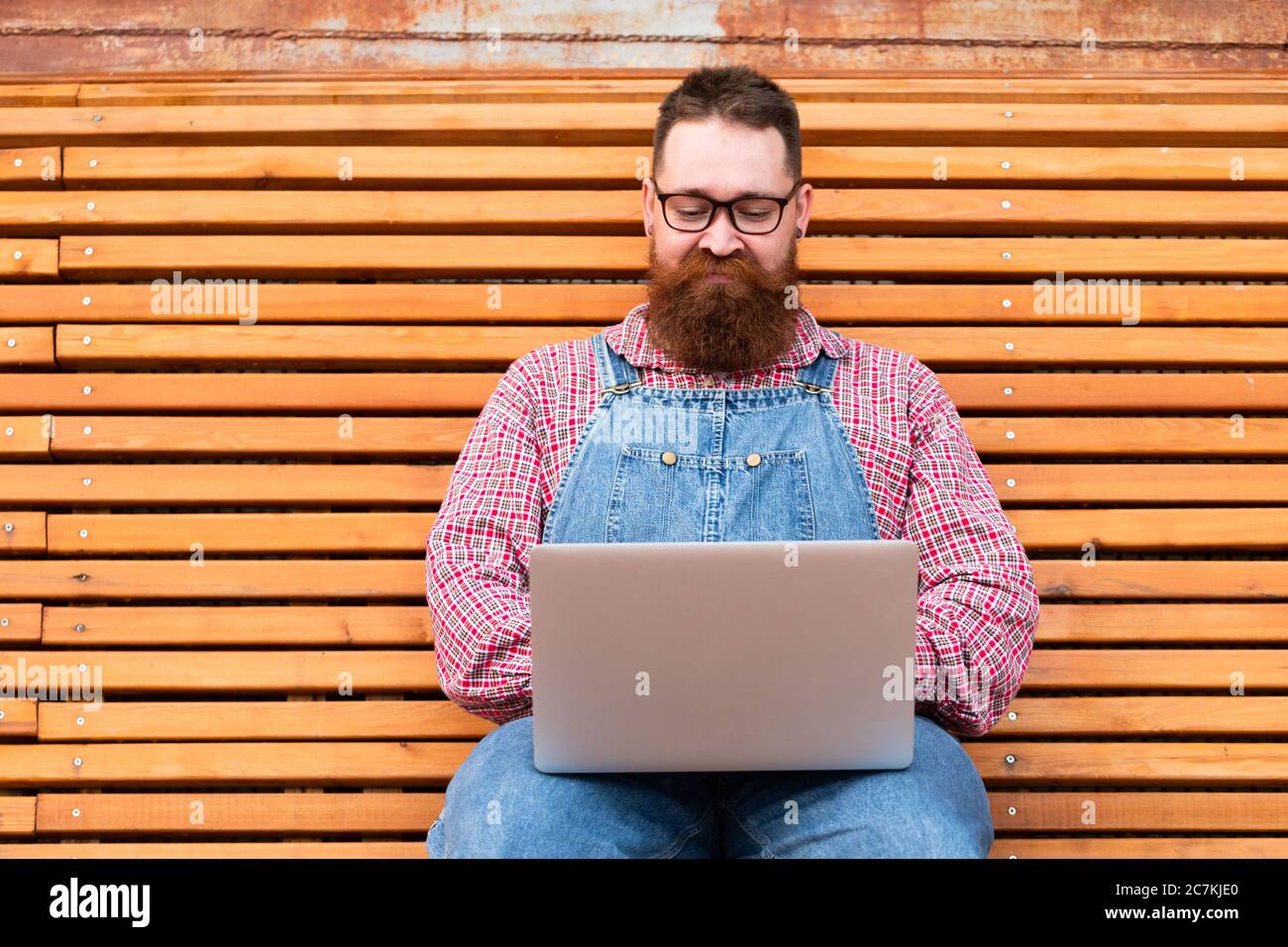 Portrait d'un boxeur hippie à barbe brutale dans une combinaison bleue, chemise à carreaux travaillant sur un ordinateur portable assis sur un banc à l'extérieur. Travail à distance, travail à distance. Banque D'Images