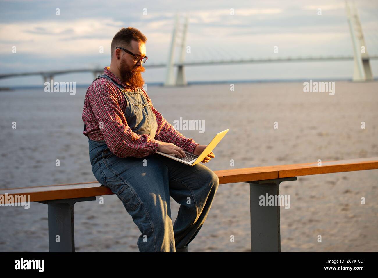 Portrait d'un homme à barbe brutal indépendant portant une combinaison bleue et une chemise à carreaux travaillant sur un ordinateur portable à l'extérieur. Travail à distance, travail à distance. Banque D'Images