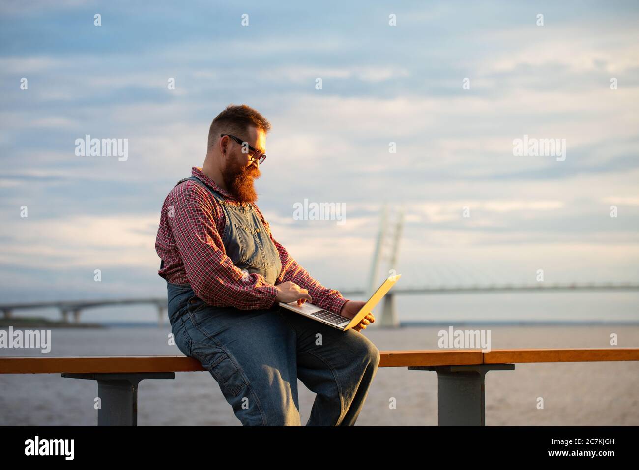 Portrait d'un homme à barbe brutal indépendant portant une combinaison bleue et une chemise à carreaux travaillant sur un ordinateur portable à l'extérieur. Travail à distance, travail à distance. Banque D'Images