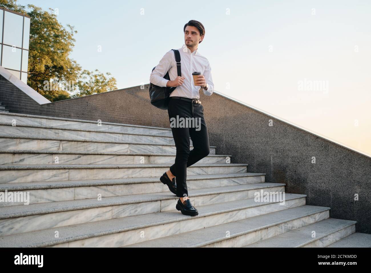 Jeune homme de brunette à barbe en chemise blanche avec écouteurs sans fil tenant une tasse de café à la main et sac à dos sur l'épaule judicieusement Banque D'Images