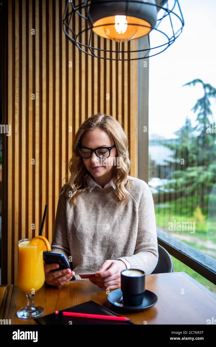 Femme blonde élégante assise au café tenant le téléphone mobile et la carte de crédit s'inscrire sur le site Web. Femme d'affaires payant avec une carte de crédit pendant ses achats Banque D'Images