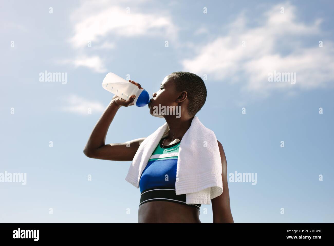 Femme de fitness buvant de l'eau contre le ciel. Une athlète féminine boit de l'eau à l'extérieur après une séance d'entraînement. Banque D'Images