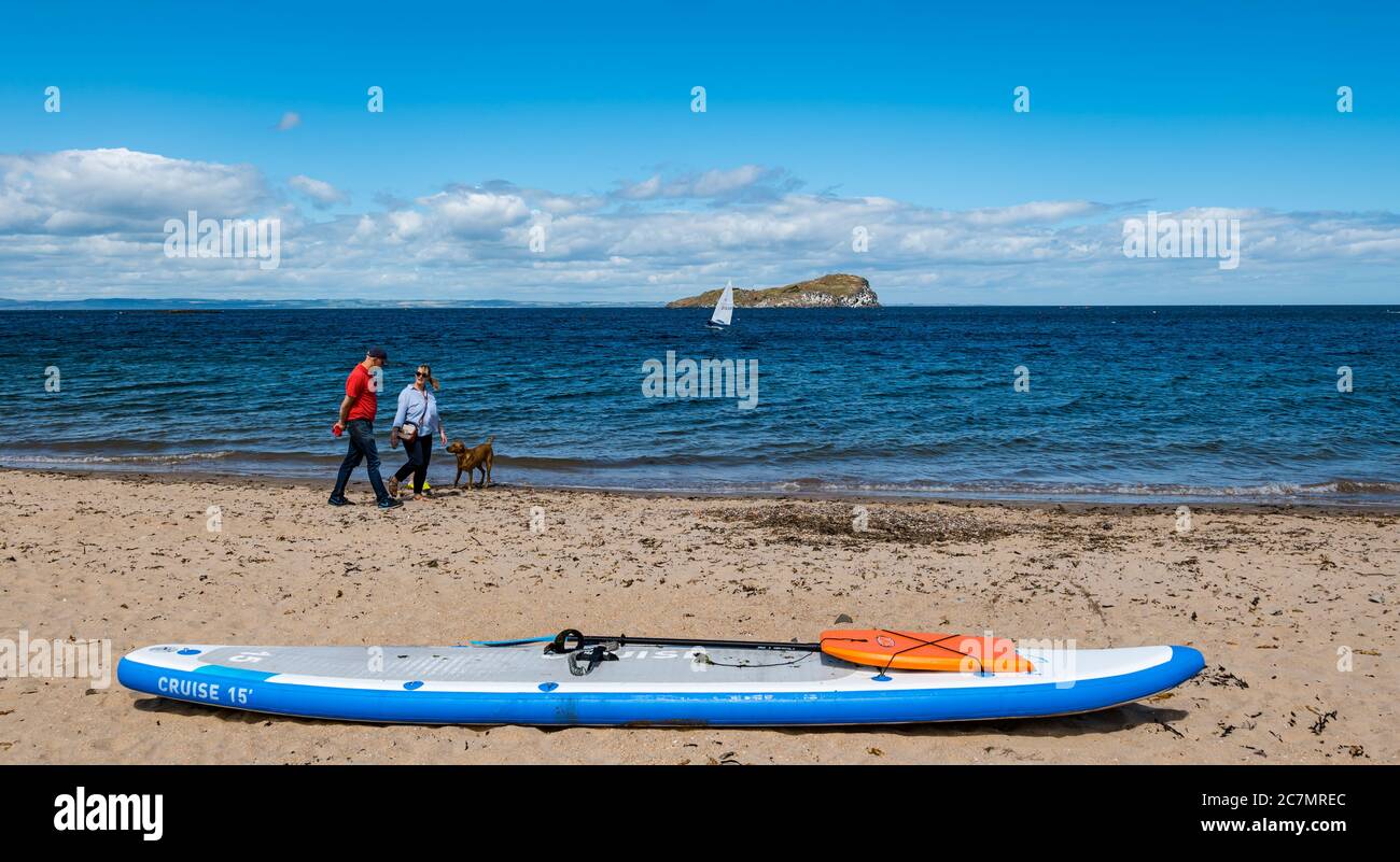 Un couple de marche sur la plage avec un paddle board dans le sable en été soleil, Berwick Nord, Lothian est, Ecosse, Royaume-Uni Banque D'Images