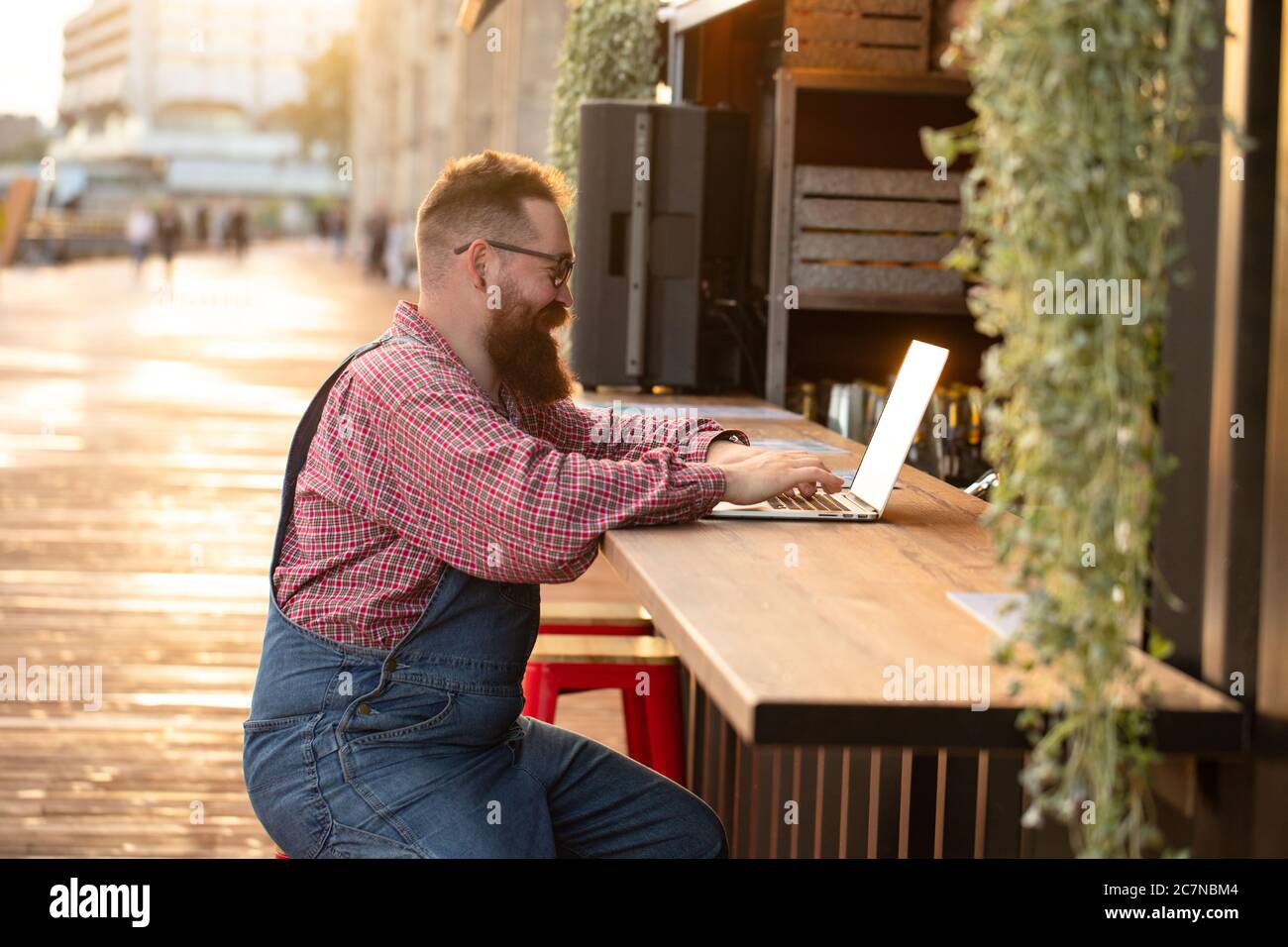 Portrait d'un boxeur hippster freelance à barbes, portant une combinaison bleue et une chemise à carreaux, travaillant sur un ordinateur portable assis dans un café/restaurant à l'extérieur. Côté Banque D'Images