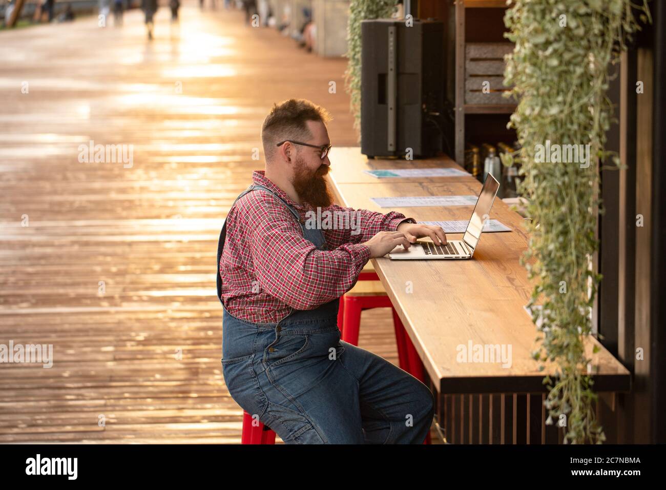 Portrait d'un boxeur hippster freelance à barbes, portant une combinaison bleue et une chemise à carreaux, travaillant sur un ordinateur portable assis dans un café/restaurant à l'extérieur. Côté Banque D'Images