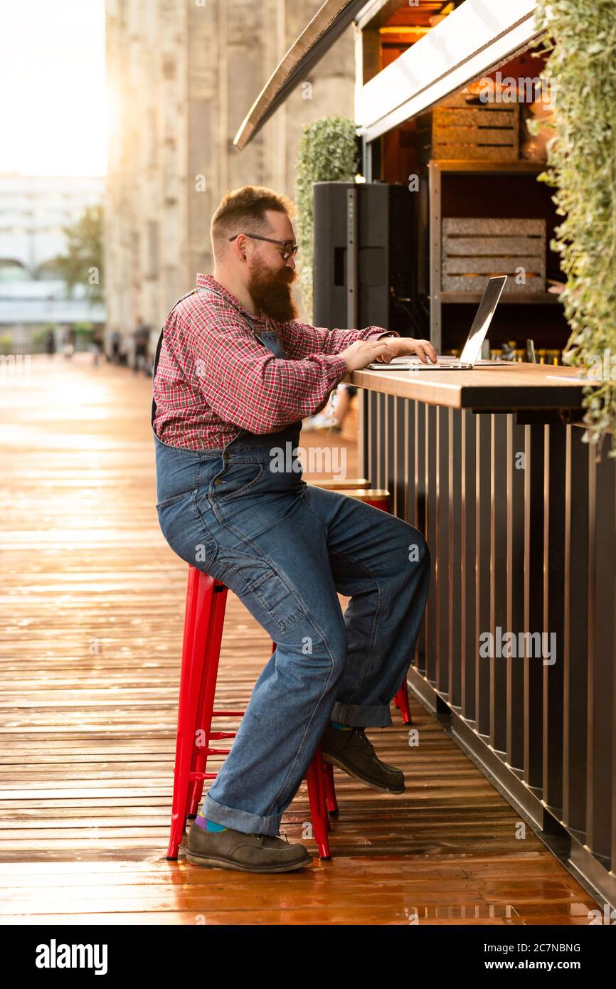 Portrait d'un boxeur hippster freelance à barbes, portant une combinaison bleue et une chemise à carreaux, travaillant sur un ordinateur portable assis dans un café/restaurant à l'extérieur. Côté Banque D'Images