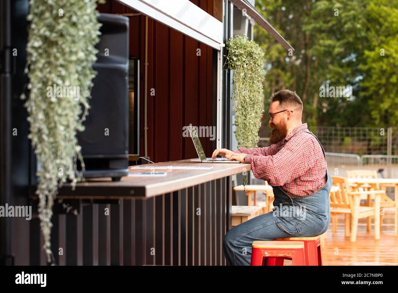 Portrait d'un boxeur hippster freelance à barbes, portant une combinaison bleue et une chemise à carreaux, travaillant sur un ordinateur portable assis dans un café/restaurant à l'extérieur. Côté Banque D'Images