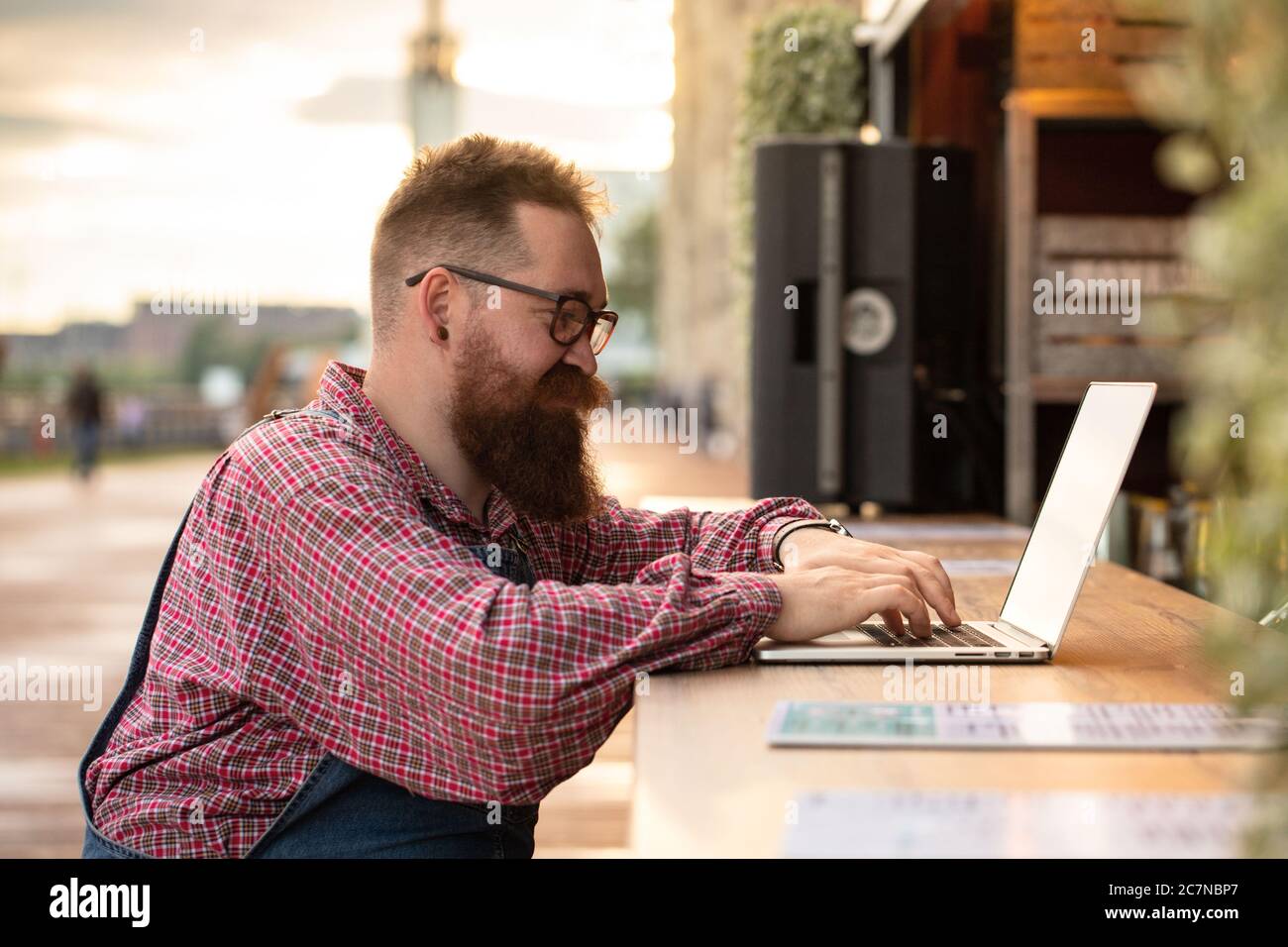 Portrait d'un boxeur hippster freelance à barbes, portant une combinaison bleue et une chemise à carreaux, travaillant sur un ordinateur portable assis dans un café/restaurant à l'extérieur. Côté Banque D'Images