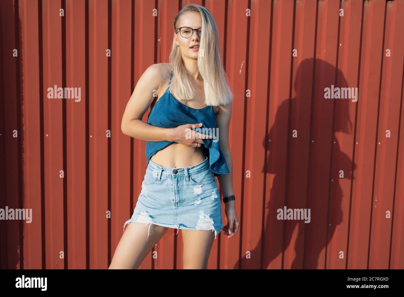 Belle jeune fille millénaire avec une coupe moderne et des lunettes posant gaiement sur un mur rouge. Elle porte une jupe de Jean et un chemisier facile pour l'été Banque D'Images