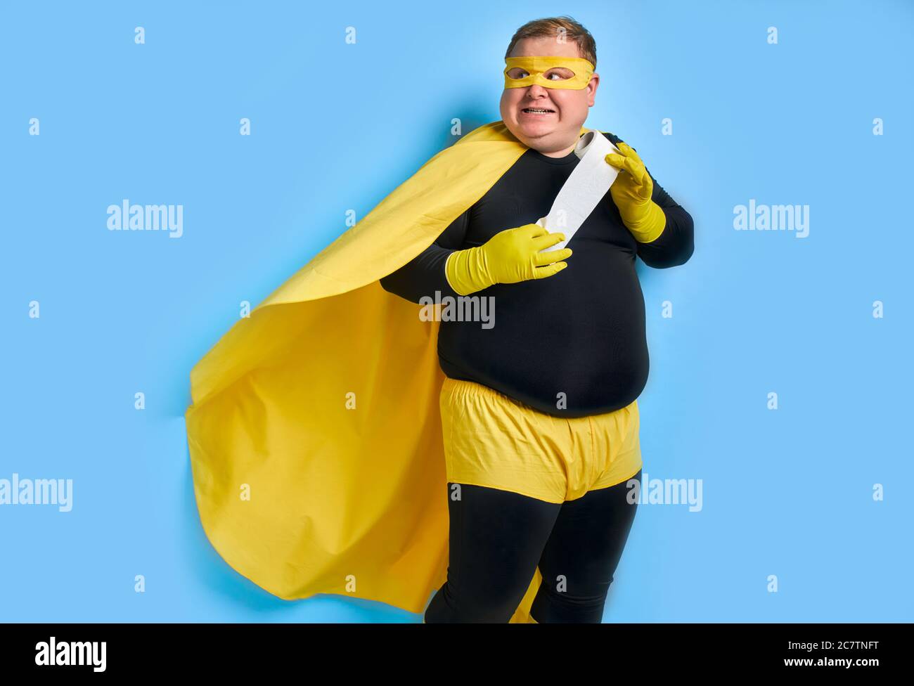 portrait de drôle caucasien gros homme avec rouleau de papier toilette dans les mains, l'homme porte le costume noir et jaune avec masque, il regarde côté avec peur f Banque D'Images