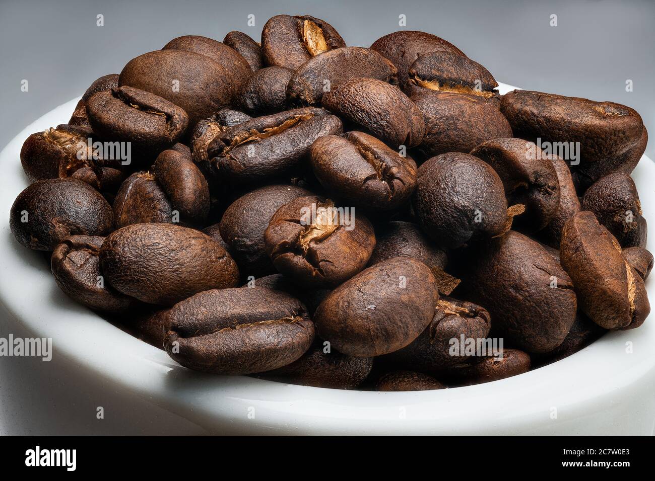 Macro de gros plan extrême d'une pile de grains de café torréfiés dans une petite tasse isolée sur un fond blanc et gris Banque D'Images