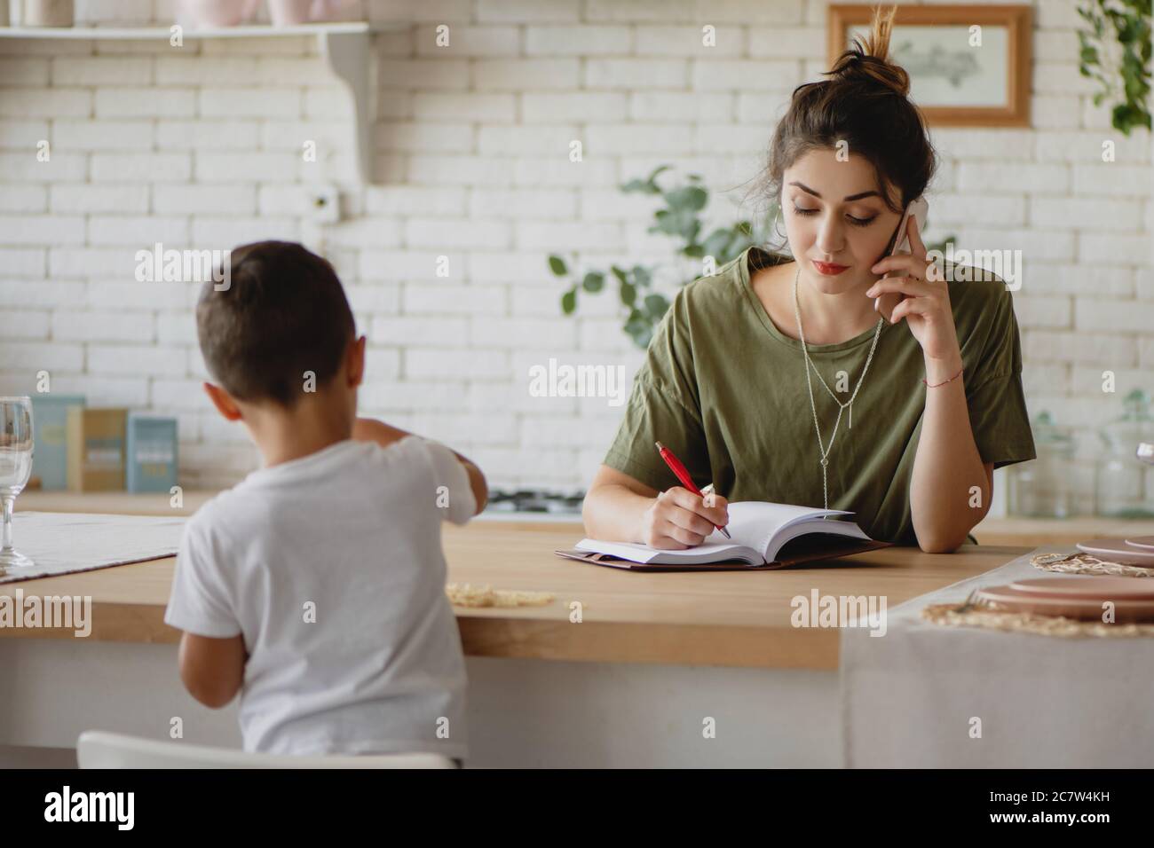 Jeune femme prenant des notes et parlant au téléphone pendant que son enfant joue à proximité. Concept de mère de travail. Banque D'Images