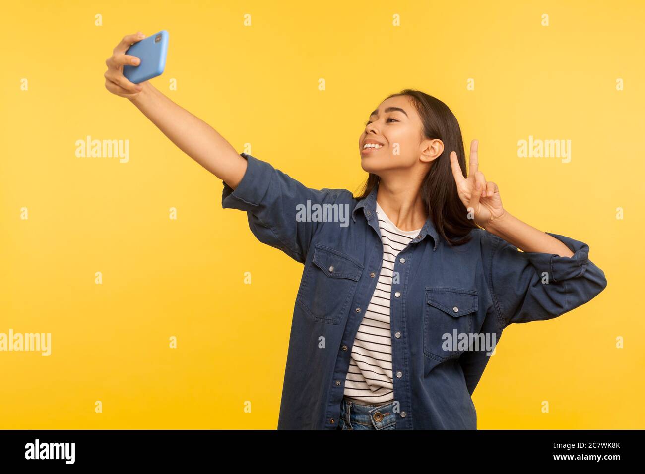 Portrait d'une fille élégante réussie en chemise denim prenant le selfie ou parlant sur appel vidéo et montrant la victoire, le geste de paix, agréable communic en ligne Banque D'Images